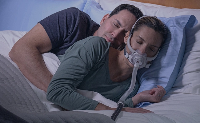 Ein Paar schläft aneinander gekuschelt im Bett, wobei die Frau eine AirFit F40 CPAP-Maske trägt.