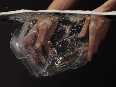 Ein Paar Hände, die in der Wasserkammer eines Luftbefeuchters eines ResMed CPAP-Geräts auf schwarzem Hintergrund eintauchen.