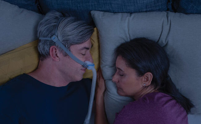Nahe bei seiner Frau schlafender Mann mit CPAP-Maske AirFit N30